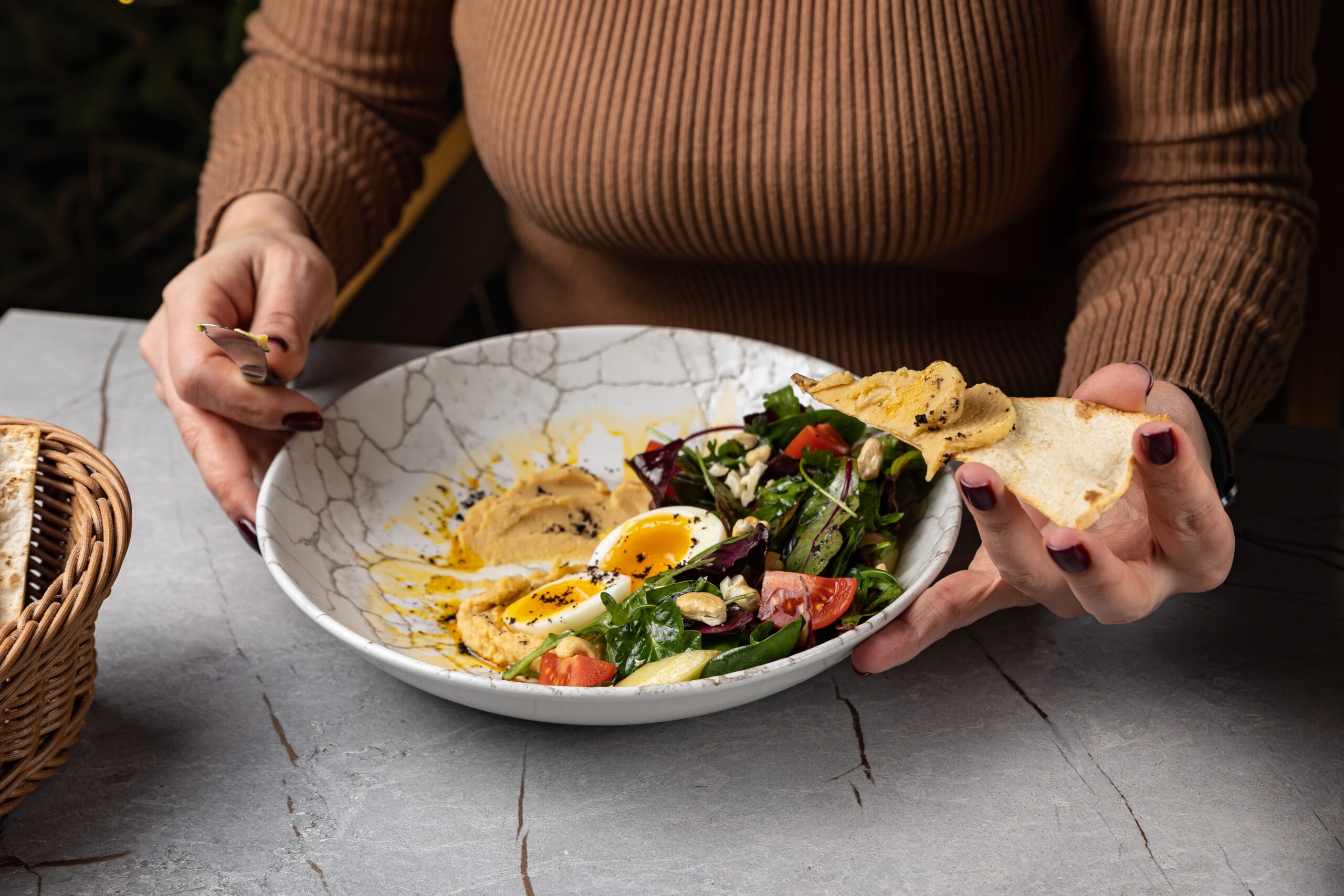 Woman spreading hummus on flatbread with gourmet salad and artisan ingredients.