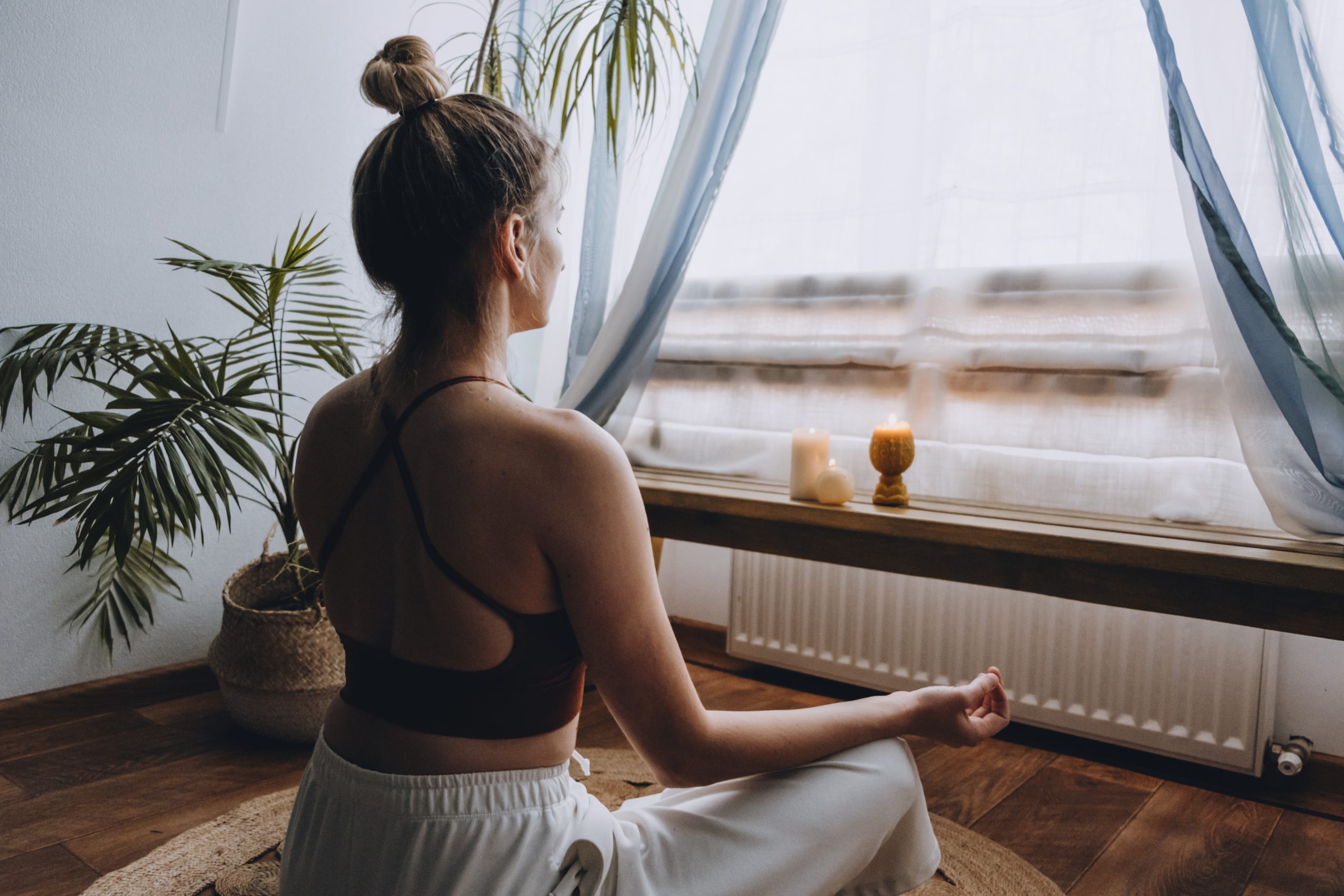 Woman meditating near window with candles and plants, promoting mental wellbeing and self-care.