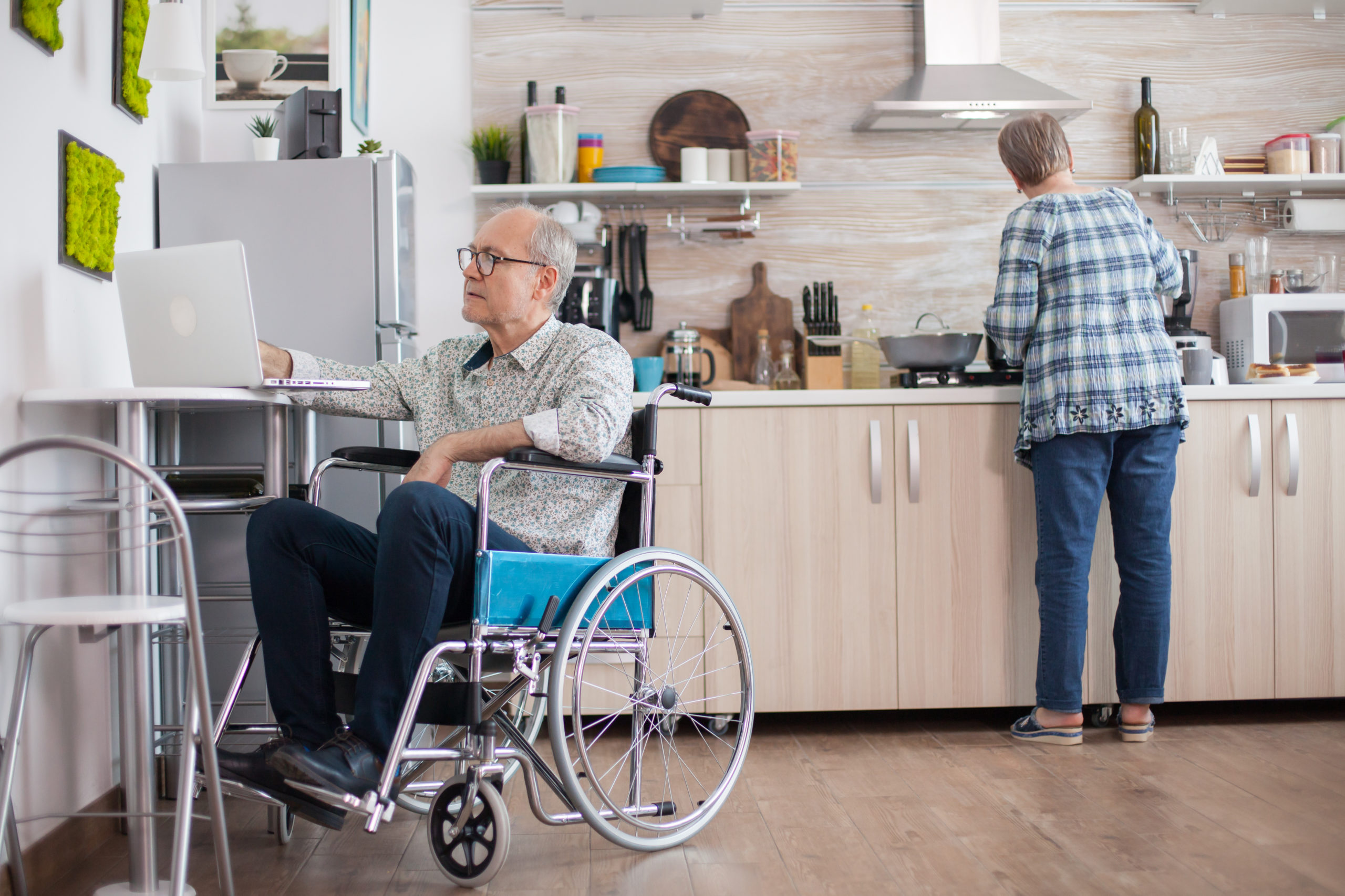 Man in wheelchair using laptop in accessible kitchen, caregiver preparing food in background.